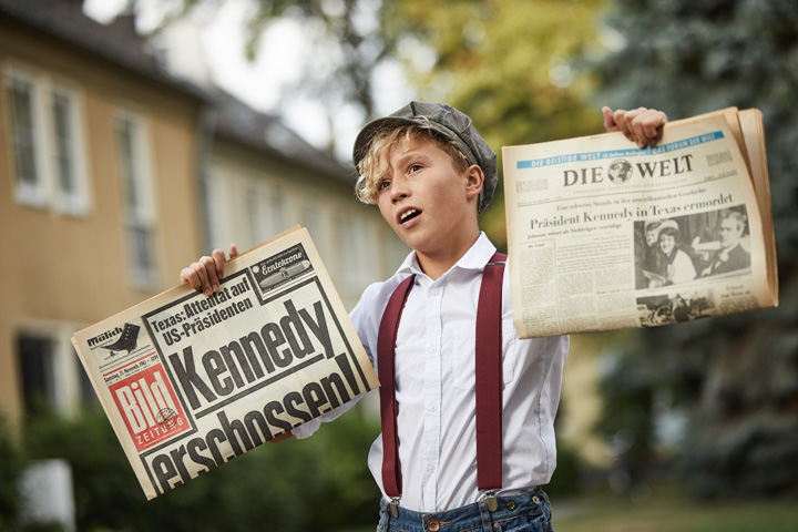 Zeitung zum Geburtstag: Man sieht einen Zeitungsjungen (Verkäufer) der eine Bild Zeitung und die Welt Zeitung , mit den Schlagzeilen von Kennedys Ermordung hoch hält.